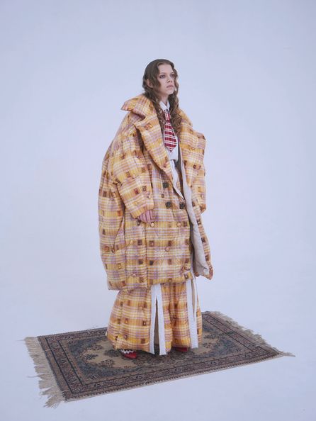 A model standing in a completely white room wearing an oversized yellow & brown tartan jacket and matching oversized pants. The model is looking off in the distance while standing on a faded persian rug.