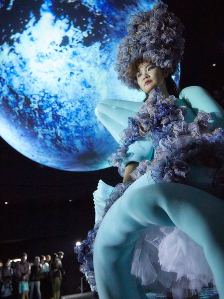 A model wearing a light blue outfit consisting of a triangular segment extending out from one shoulder, and a round doughnut-like component at the bottom. The space between the light blue segments consists of bunched, ruffled thin fabric in various shades of blue, and a matching large hat extending upwards. The model is looking to the side while standing in a dark room with a large blue-tinted moon in the background.