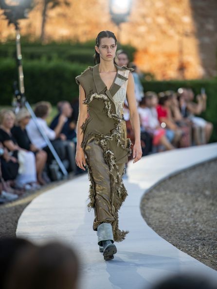A model walking along a circular catwalk wearing an outfit consisting of parts of an olive green jacket and lighter components with distressed seams where each component connects.